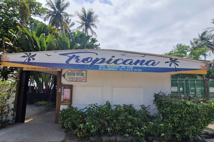 Entrée actuelle de l’Hôtel Tropicana à Libreville : bâtiment blanc sous l’enseigne Tropicana (hôtel, restaurant, bar, plage), entouré de cocotiers et de massifs fleuris